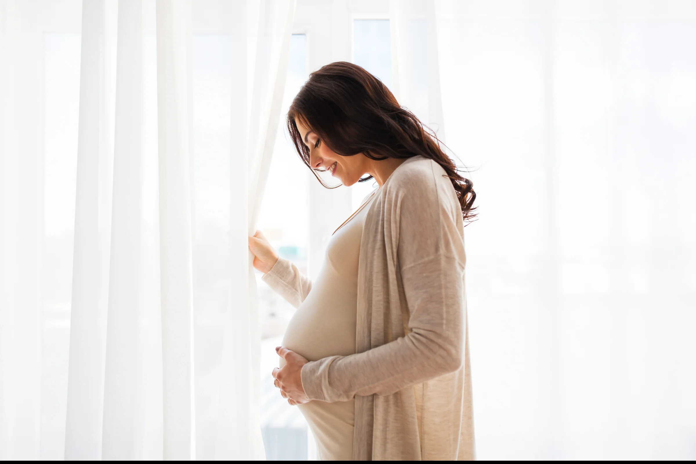 A pregnant woman at a sunlit window, one hand resting on her bump