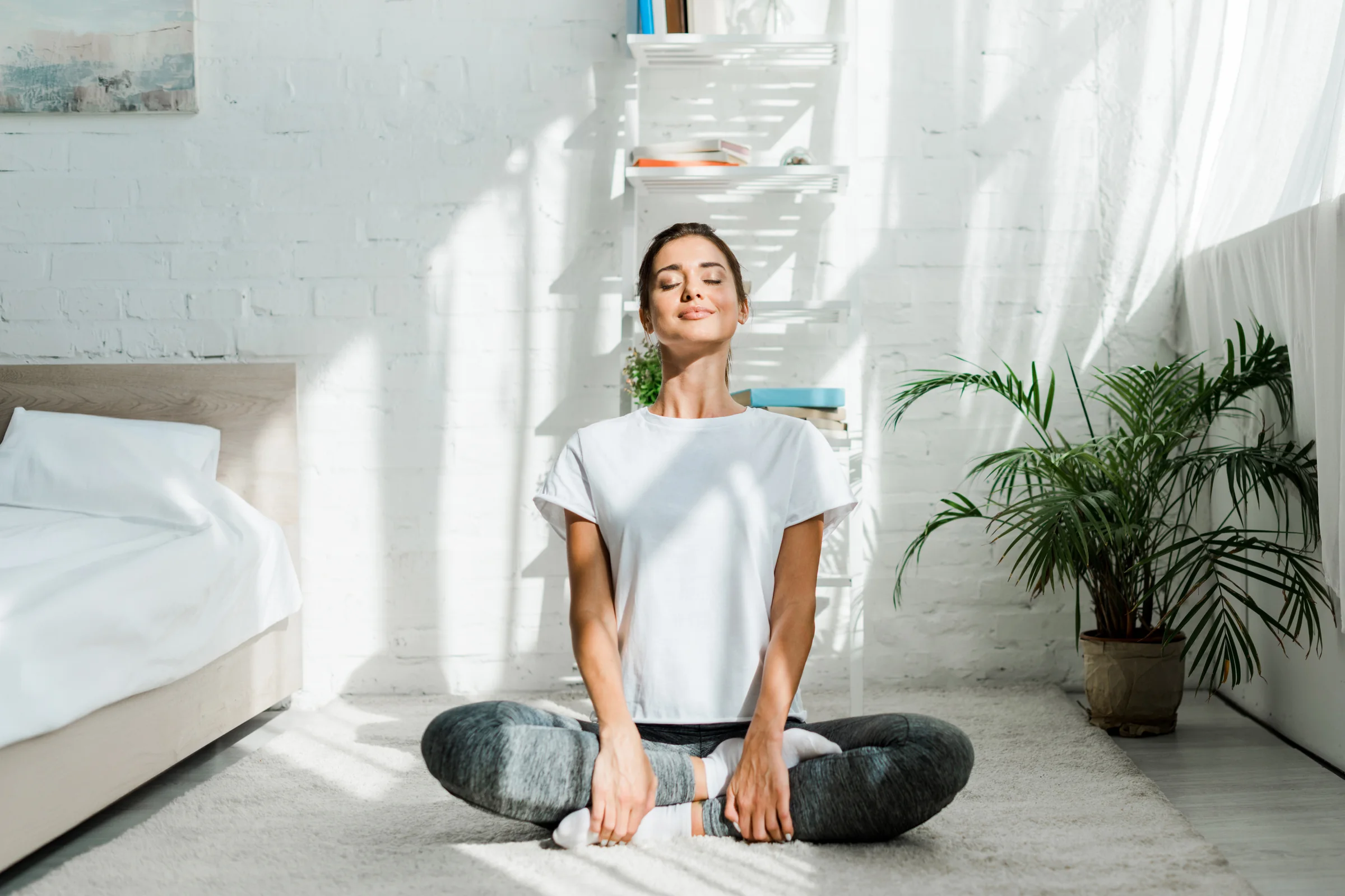 A woman sitting cross legged on her bedroom floor in morning light